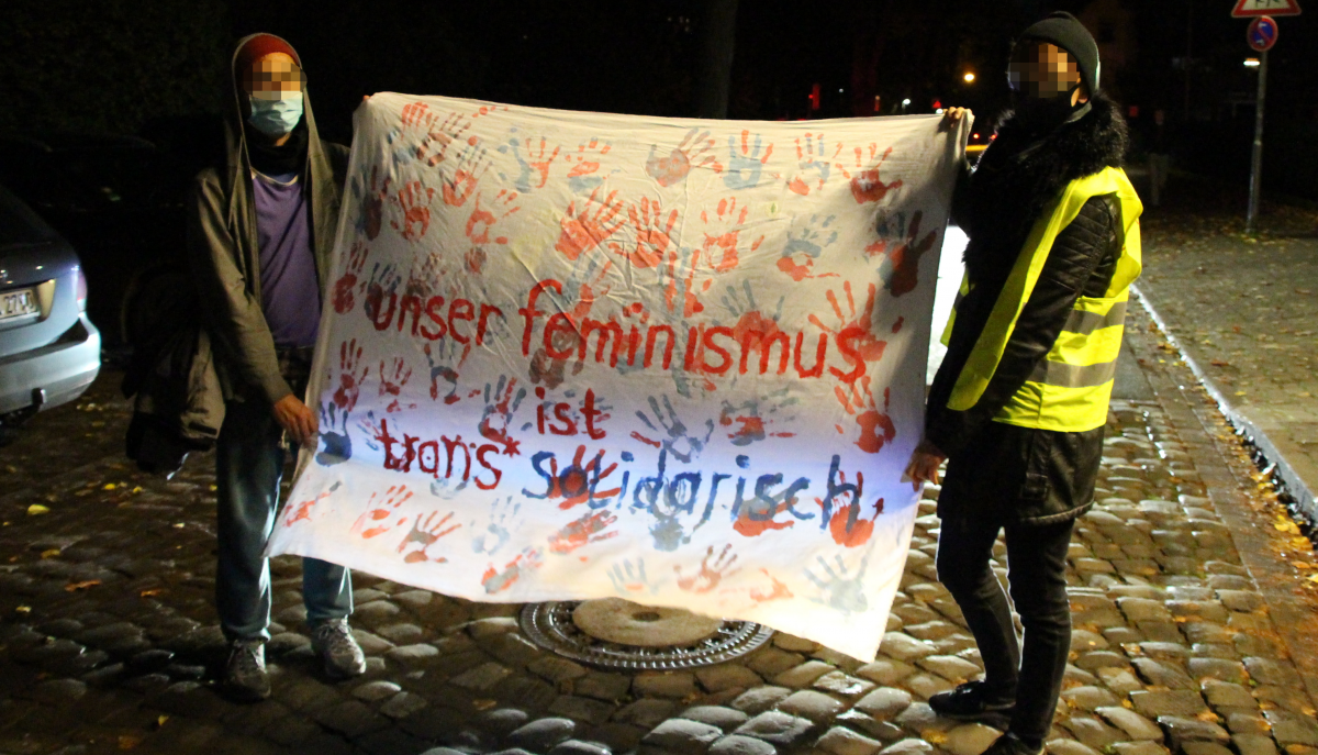 zwei menschen halten ein banner auf dem handabdrücke in den farben der transflagge zu sehen sind. darauf steht "unser feminismus ist transsolidarisch"